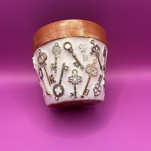 May include: A white flower pot with a gold rim, decorated with bronze and silver antique-style keys. The keys vary in shape and size, giving the pot a vintage appearance.