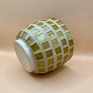 May include: A decorative vase with a mosaic tile design. The vase has a white base and a rounded body covered in square green and iridescent tiles. The vase is tilted, showing the mosaic pattern and white base.