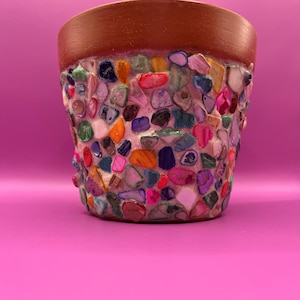 May include: A small, handcrafted flower pot with a brown rim and a body encrusted with colourful, polished stones. The stones are various shapes and sizes, including red, blue, green, and purple, creating a mosaic-like texture. The pot sits on a pink background.
