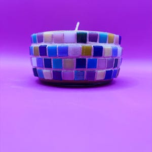 May include: A mosaic candle holder with a white wick. The candle holder is cylindrical and features a mosaic design with small, square tiles in shades of blue, purple, yellow, and white. The background is a solid purple.