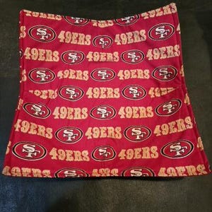 May include: A red and gold patterned fabric item featuring the text "49ERS" and the team logo. The design is repeated across the fabric. The item appears to be a pillowcase or similar fabric product.