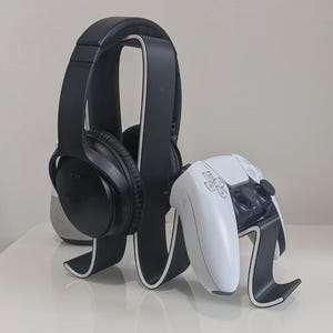 May include: Black over-ear headphones rest on a black and white headphone stand next to a white and black game controller on a white surface. The stand has a curved design, and the controller is also on a stand.