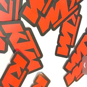May include: A collection of orange and black KTM logo stickers. The KTM logo is a stylized design with the letters "K" and "M" in a bold, angular font. The stickers are scattered against a white background.