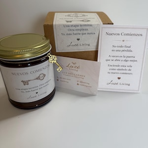May include: A candle in a glass jar with a gold lid, a small gold key charm, and a gift box. The label reads "Nuevos Comienzos" and includes text in Spanish. A business card is also present. The candle is a dark amber color.