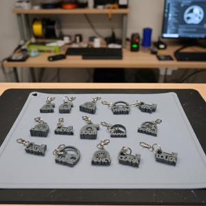 Peut inclure: Ensemble de porte-clés gris imprimés en 3D, avec des noms et des motifs variés. Chaque porte-clé est équipé d'un fermoir argenté. Les porte-clés sont disposés sur un tapis gris, avec un bureau et un ordinateur en arrière-plan.