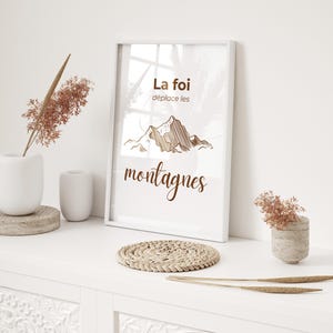 Peut inclure: Un cadre blanc avec l'inscription "La foi déplace les montagnes" et une illustration de montagne marron. Il est posé sur une surface blanche avec des éléments décoratifs, dont un dessous de verre tressé et des plantes séchées.