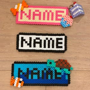 May include: Three handmade name tags made from colorful plastic beads. The top tag is pink and yellow with the word "NAME" in pink. The second tag is black and white with the word "NAME" in white. The bottom tag is blue and teal with the word "NAME" in teal.