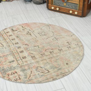 May include: A round area rug featuring a faded, distressed pattern in muted shades of beige, soft pink, and sage green. The design includes intricate geometric borders and central motifs with a weathered, vintage-inspired appearance.