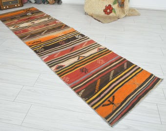 May include: A long, narrow runner rug featuring horizontal stripes in shades of orange, red, brown, yellow, and cream. The rug is decorated with small, repeating geometric patterns and stylized motifs distributed across the striped surface.