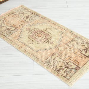 May include: Rectangular, vintage-style rug with a central geometric design in shades of beige, brown, and peach. The rug has a fringed edge and is placed on a light-colored wooden floor. The rug's pattern includes repeating geometric shapes.