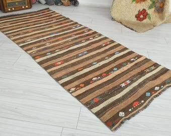 May include: A rectangular woven runner rug featuring horizontal stripes in various shades of brown, tan, and cream. The rug is decorated with small, scattered geometric motifs in red, blue, green, and white, and has a fringed edge on both ends.