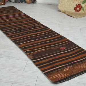 May include: A long, narrow runner rug featuring a series of horizontal stripes in shades of dark brown, black, and copper. The textile includes subtle geometric patterns and thin, intermittent lines of pink and orange woven throughout the fabric.