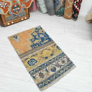 May include: A small, rectangular rug with an intricate pattern. The rug features a combination of orange, blue, and yellow hues. The design includes floral and geometric elements, creating a vintage or bohemian aesthetic. The rug is likely made of woven fabric.