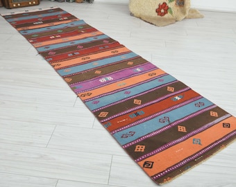 May include: A long, narrow runner rug featuring horizontal stripes in shades of brown, orange, light blue, and purple. The rug is decorated with small, repeating geometric patterns in contrasting colors, distributed evenly across the entire surface.