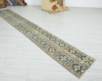 May include: A long, narrow runner rug featuring a repeating geometric pattern in shades of dark blue, muted yellow, and light blue. The design consists of interconnected square motifs and linear borders, creating a structured, symmetrical appearance.