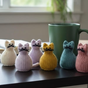May include: A collection of six knitted cat figurines in various colors, including cream, lavender, yellow, teal, and pink. Each cat has black whiskers and a small tail. A green mug and an open book are in the background.
