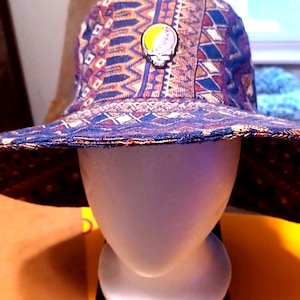 May include: Reversible bucket hat with a colorful geometric pattern in blue, red, and white. The hat features a skull and lightning bolt pin on the front. The hat is displayed on a white mannequin head.