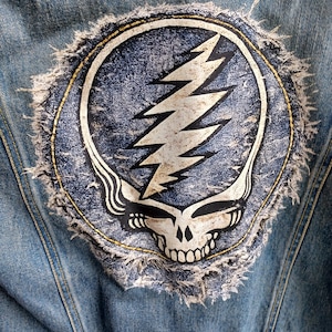 May include: A light blue denim jacket with a distressed Steal Your Face patch on the back. The patch features a white skull and lightning bolt design on a blue background, with a frayed edge.