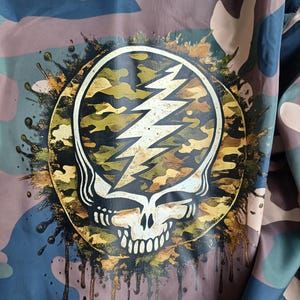 May include: Camouflage-patterned fabric with a large, circular graphic. The graphic features a white skull with a lightning bolt, surrounded by a gold and camouflage design. Gold paint drips from the graphic onto the fabric.
