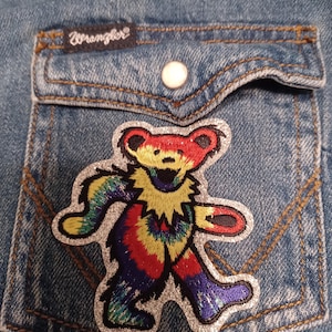 Könnte beinhalten: Nahaufnahme eines hellblauen Jeanshemdes mit einer Tasche. Ein farbenfroher, gestickter Bärenaufnäher mit Batik-Design befindet sich auf der Tasche. Das Wrangler-Logo ist über der Tasche sichtbar.