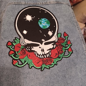 May include: Light blue denim jacket featuring a large patch on the back. The patch depicts a skull with a starry sky and a blue and green planet inside. Red roses and green leaves surround the skull, creating a vibrant contrast.
