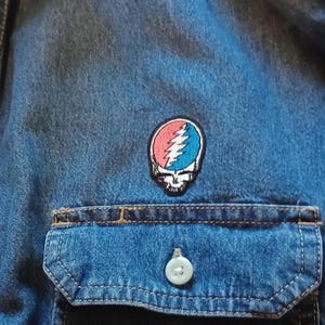 May include: Close-up of a blue denim shirt with a pocket and a white button. A colorful patch featuring a skull with a lightning bolt design in red and blue is sewn on the shirt.