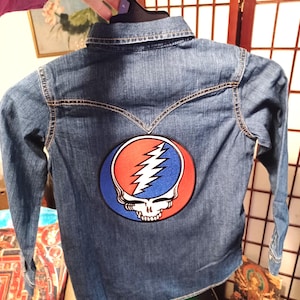 May include: Blue denim shirt with long sleeves and a collar. The back of the shirt features a large, circular patch with a skull and lightning bolt design in red, white, and blue. The shirt is hanging on a black hanger.