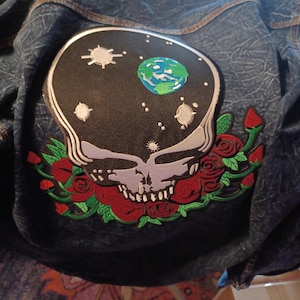 May include: Dark blue denim jacket with a large embroidered patch featuring a skull with a starry sky design and a green and blue Earth. Red roses and green leaves surround the skull.