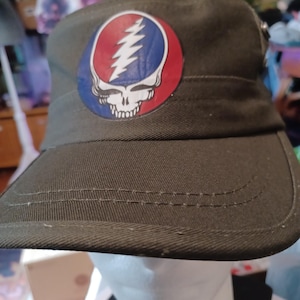 May include: Olive green hat with a brim and a round patch featuring a skull with a lightning bolt and a red, white, and blue color scheme. The hat is made of a durable fabric and has a structured design.