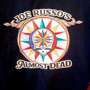 May include: Black t-shirt featuring a colorful graphic. The graphic includes a skull, a starburst design, and the text "JOE RUSSO'S ALMOST DEAD" in a stylized font. The design incorporates red, blue, yellow, and teal.