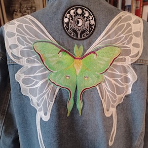 May include: A denim jacket with a large green and red moth applique on the back. White lace-like wings are sewn on the shoulders. A circular patch with a moon phase design is above the moth.