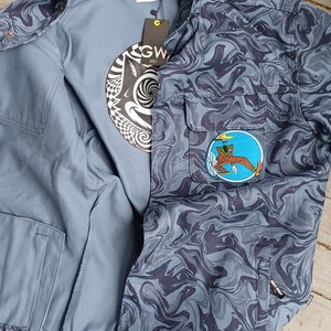 May include: A blue jacket with a swirling dark blue pattern, featuring a patch with a cartoon figure. The jacket is open, revealing a gray t-shirt with a black and white graphic. The jacket has two front pockets and a tag.