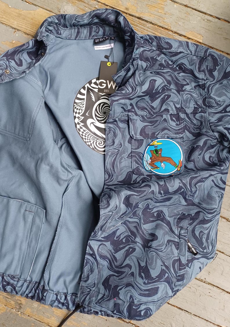 May include: A blue jacket with a swirling dark blue pattern, featuring a patch with a cartoon figure. The jacket is open, revealing a gray t-shirt with a black and white graphic. The jacket has two front pockets and a tag.