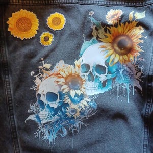 May include: Dark denim jacket with a floral and skull design. The back of the jacket features two skulls with sunflowers and blue floral accents. Additional sunflower patches are scattered across the jacket. A pin with a lightning bolt design is at the top.