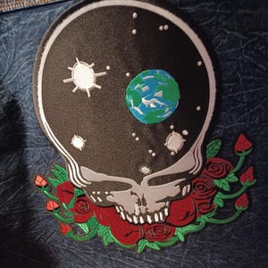 May include: Embroidered patch featuring a skull with a starry night sky design, including a planet Earth. The skull is surrounded by red roses and green leaves. The patch is attached to a denim jacket.