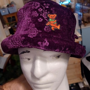 May include: A deep purple velvet bucket hat with floral embroidery and a small orange bear patch. The hat has a rounded crown and a slightly curved brim. The bear patch is wearing a green scarf.