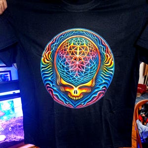 May include: Black t-shirt featuring a vibrant, psychedelic skull design. The skull is rendered in a spectrum of colors, including blue, yellow, pink, and orange, with intricate geometric patterns. The design is centered on the shirt.