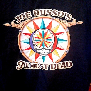 May include: Black t-shirt featuring a colorful graphic design. The design includes a circular emblem with a skull, starburst, and the text "JOE RUSSO'S ALMOST DEAD". The emblem is surrounded by a decorative border.