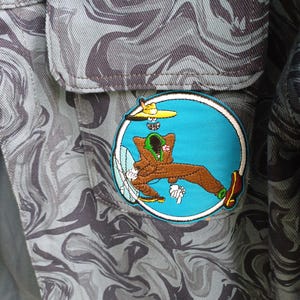May include: Close-up of a gray and black camouflage patterned fabric with a patch. The patch features a cartoon character in a blue circle with a yellow hat and brown outfit. The fabric has a pocket.