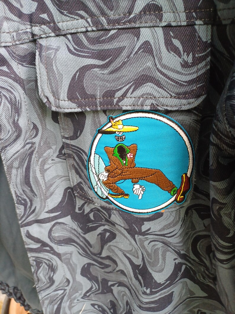 May include: Close-up of a gray and black camouflage patterned fabric with a patch. The patch features a cartoon character in a blue circle with a yellow hat and brown outfit. The fabric has a pocket.