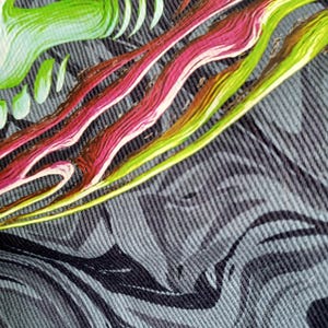 May include: Close-up of a textile with a swirling, abstract design. The pattern features flowing lines in shades of green, pink, and yellow against a textured gray background. The fabric appears to be denim or a similar material.