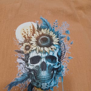 May include: A tan-colored shirt featuring a detailed graphic of a skull adorned with sunflowers and blue foliage. The skull has a teal hue, with intricate shading and floral accents. The design includes a full moon and dripping water effects.