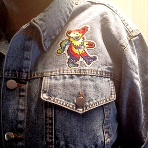 May include: A denim jacket with a colorful bear patch on the upper left chest. The patch features a dancing bear design in shades of red, yellow, blue, and green. The jacket has silver buttons and a classic pocket design.