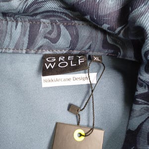 May include: Close-up of a denim jacket with a blue and navy leaf pattern. The jacket has a collar and a tag that reads "GREY WOLF" and "NikkiArcane Design". The size is XL.
