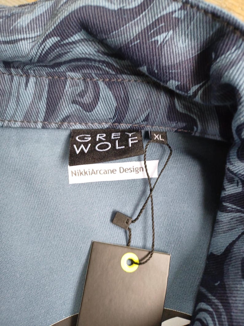 May include: Close-up of a denim jacket with a blue and navy leaf pattern. The jacket has a collar and a tag that reads "GREY WOLF" and "NikkiArcane Design". The size is XL.