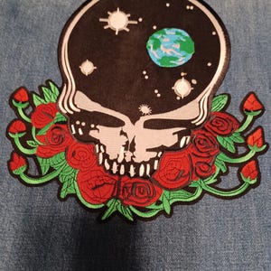 Puede incluir: Parche bordado con una calavera sobre un fondo negro que representa un cielo estrellado y un planeta azul y verde. La calavera está rodeada de rosas rojas con hojas verdes, sobre un fondo vaquero.