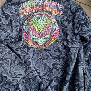 May include: A dark blue jacket with a swirling pattern and a colorful skull graphic on the back. The text "Grateful Dead" is arched above the skull. The jacket has a zipper and a drawstring at the bottom.