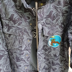 May include: A gray and black patterned jacket with two chest pockets and a button-up front. The jacket features a cartoon patch on the right side. The pattern resembles a swirling marble design.