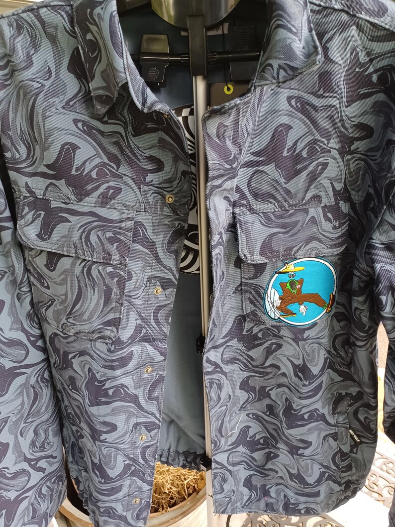 May include: A gray and black patterned jacket with two chest pockets and a button-up front. The jacket features a cartoon patch on the right side. The pattern resembles a swirling marble design.