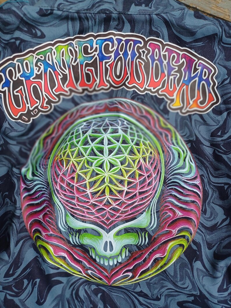 May include: A close-up of a shirt with a colorful skull design. The skull is surrounded by a vibrant, geometric pattern in shades of green, pink, and yellow. The text "Grateful Dead" is arched above the skull in a rainbow of colors against a dark, swirling background.